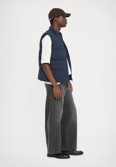 Vesta umplută de culoare bleumarin cu guler înalt, purtată peste o cămașă albă, asociată cu o pereche de pantaloni largi din denim gri și cizme negre, văzută din lateral.