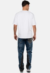 T-shirt en coton blanc à manches courtes, associé à un jean bleu avec des panneaux contrastants noirs et camouflage, vue de dos.
