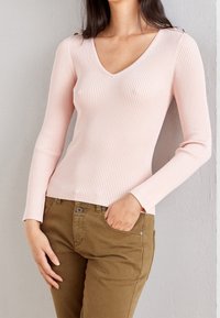 Una donna indossa un maglione a maniche lunghe rosa chiaro a costine con scollatura a V e pantaloni marroni, in piedi con una mano nella tasca.