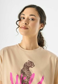 T-shirt en coton beige avec un graphique de léopard marron et le texte "Wild" en rose. Design à col rond avec des manches courtes et texture lisse.