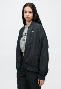 Μαύρο αθλητικό μπουφάν με κούμπωμα με κουμπιά και ριμπ τελειώματα στα μανίκια. Φορεμένο πάνω από γκρι κοντομάνικο φούτερ με μαύρο κείμενο.