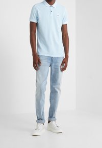 Světle modrá polo tričko s krátkými rukávy a malým logem na hrudi, spárované se světle vypranými džíny a bílými teniskami. Neformální střih a jednoduchý design.