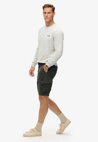 Homme marchant, portant une chemise à manches longues gris clair, un short cargo vert foncé, des chaussettes beiges et des baskets beiges, sur un fond blanc uni.