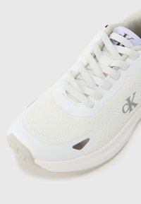 Λευκό αθλητικό παπούτσι με πλέγμα στο πάνω μέρος, υφή στην λεπτομέρεια, ενισχυμένη μύτη και λογότυπο. Διαθέτει δέσιμο με κορδόνια και ενισχυμένη σόλα για άνεση.