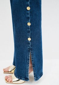 Jeans de denim de talle alto en azul oscuro con pierna recta, que cuentan con una abertura en el dobladillo y botones en tono dorado a lo largo de la costura lateral.