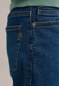 Jeans de mezclilla azul oscuro con una textura desgastada, que presentan un pequeño logo bordado en el bolsillo trasero y costuras amarillas en contraste.