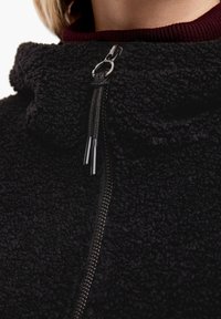 Giacca in pile nera con colletto alto, dotata di zip con accessori in metallo e tessuto testurizzato. Design caldo e casual.