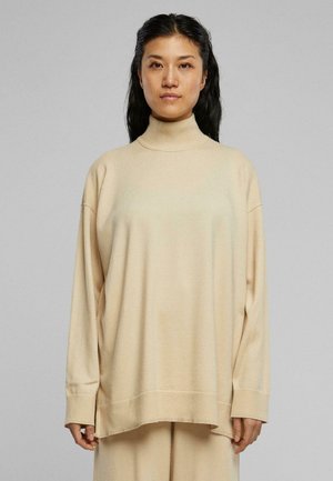 Beige polokragejumper gjord av ett mjukt material; har en lös passform, långa ärmar och ribbade mudd och fåll för extra textur.