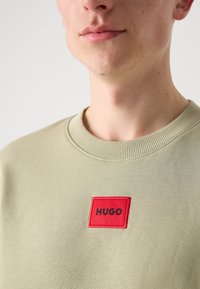 Sweatshirt vert olive clair avec un col côtelé et un patch rectangulaire rouge bien visible affichant le mot "HUGO" en texte noir.