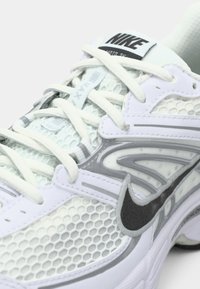 Λευκό και γκρι αθλητικό παπούτσι της Nike με μαύρο λογότυπο Swoosh, πλέγμα επάνω μέρος και λευκές κορδόνες δεμένες πάνω στη γλώσσα.