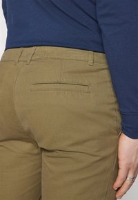 Khaki kalhoty s rovnými nohavicemi. Vyrobené z bavlny, mají zadní kapsu a lehkou, hladkou texturu. Nosené s námořnickou modrou košili s dlouhým rukávem.