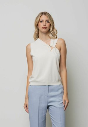 Donna bionda con top bianco senza maniche con scollatura asimmetrica e pantaloni azzurri, in piedi contro uno sfondo semplice.