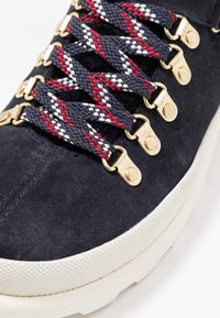 Navy suede sneaker featuring gold hardware, textured white sole, and multicolored braided laces in red, white, and navy.