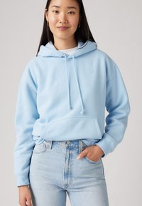Felpa azzurra chiara con tasca a marsupio, cordini e un piccolo logo del brand; abbinata a jeans azzurri chiari a vita alta.