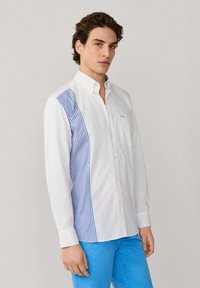 Jonge man in een wit overhemd met knopen, met blauwe verticale strepen op de zijpanelen, en felblauwe broek, staand tegen een effen achtergrond.