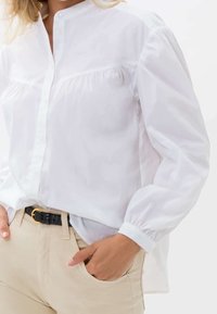Camisa blanca de botones con cuello mao y detalles fruncidos, combinada con pantalones caqui y un cinturón negro con hebilla dorada.