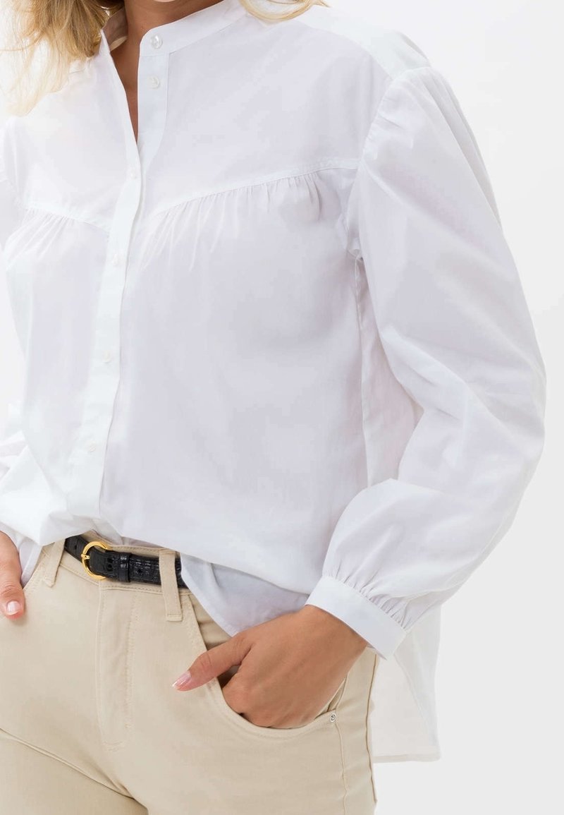 Camisa blanca de botones con cuello mao y detalles fruncidos, combinada con pantalones caqui y un cinturón negro con hebilla dorada.