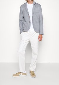 Hombre con blazer gris, camiseta blanca, pantalones blancos y zapatillas beige, de pie contra un fondo blanco.