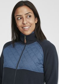 Sweatshirt en polaire marine avec un panneau en forme de diamant de couleur bleu clair sur la poitrine, col haut et fermeture éclair avec une tirette en cuir.