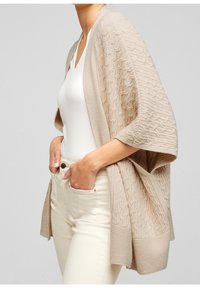 Cardigan beige a maglia con motivo testurizzato e maniche larghe. Presenta un bordo inferiore uniforme ed è indossato su una maglietta bianca e pantaloni chiari.