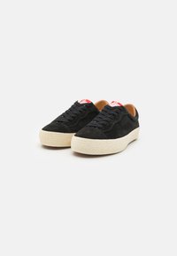 Schwarze Wildleder-Sneaker mit einer strukturierten cremefarbenen Sohle, ausgestattet mit schwarzen Schnürsenkeln und einem roten Logo-Tag an der Ferse. Minimalistisches Design, runde Form.