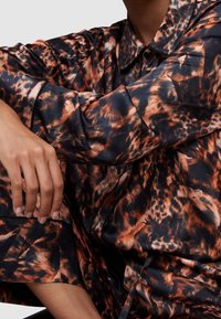 Personne portant une chemise brillante à manches longues avec un imprimé animal abstrait orange et noir, assise les bras croisés sur un genou plié.