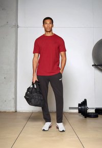 Rød kortermet t-skjorte med en liten logo, grå joggebukser, hvite sneakers med flerfargede aksenter, og en svart treningsbag med håndtak.