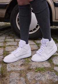 Białe skórzane sneakersy za kostkę z perforowaną czubkiem, szczegółowym przeszyciem i płaskimi sznurowadłami, w połączeniu z szarymi skarpetami do kolan na brukowanej ulicy.