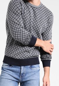 Pull à motifs bleu marine et gris avec un design en grille, poignets côtelés et col rond ; porté avec un jean en denim clair.