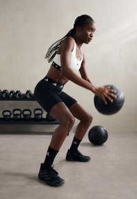 Pantaloni sportivi neri e un reggiseno sportivo bianco con una fascia nera che presenta loghi bianchi. Il soggetto tiene in mano un pallone medicinali nero, mentre è impegnato in un esercizio.