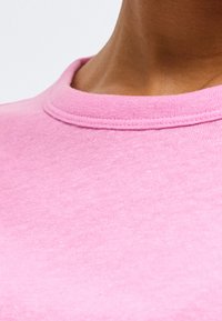 Κοντινό πλάνο στον λαιμό και το πάνω μέρος του στήθους ενός ατόμου που φοράει ροζ μπλουζάκι με στρογγυλή λαιμόκοψη σε απλό φωτεινό φόντο.