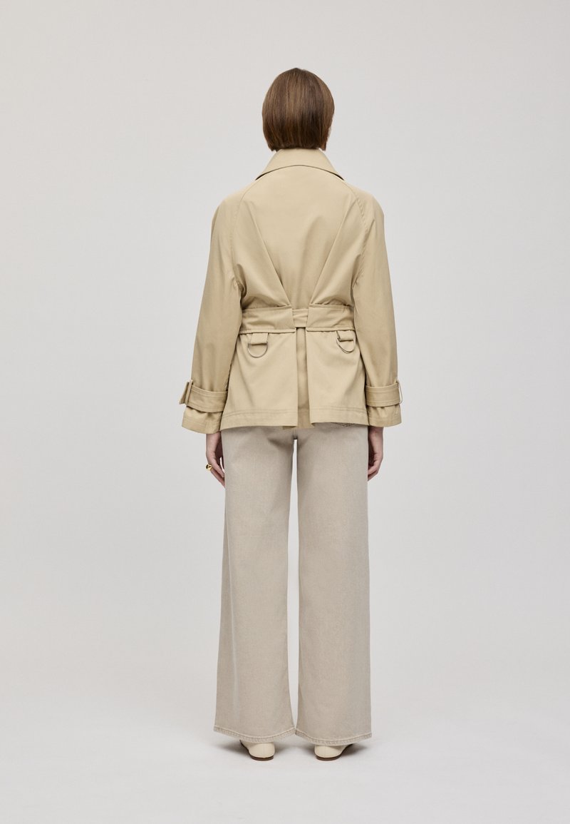 Beiger Trenchcoat mit taillierter Passform, tailliertem Gürtel, langen Ärmeln und weit ausgestellten hellbeigen Hosen. Rückansicht.
