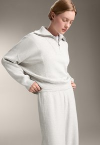Ljusgrå zip-up tröja med ribbad textur på ärmarna och kragen, ihop med matchande ljusgrå joggerbyxor.