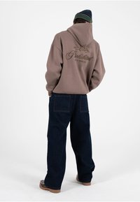 Sudadera marrón con capucha y diseño bordado en la espalda, combinada con pantalones de mezclilla azul oscuro de pierna ancha y zapatos marrones sin cordones.