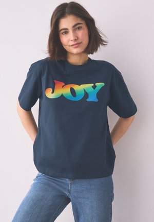 Junge Frau mit kurzen dunklen Haaren, die ein dunkelblaues T-Shirt mit dem Wort "JOY" in Regenbogenfarben und blaue Jeans trägt, stehend mit den Händen hinter dem Rücken.