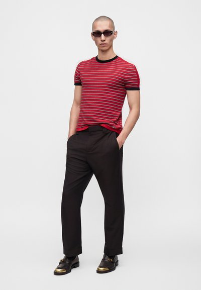 Homme portant une chemise à manches courtes rayée rouge et noire, un pantalon noir, des mocassins noirs avec des bouts dorés et des lunettes de soleil foncées, debout les mains dans les poches.