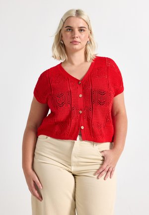 Giovane donna con capelli biondi che indossa un cardigan rosso a maniche corte in maglia, abbottonato sopra pantaloni beige, in piedi davanti a uno sfondo bianco.