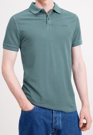 Homme portant un polo vert à manches courtes avec col et un jean en denim bleu, une main dans la poche, sur fond blanc uni.