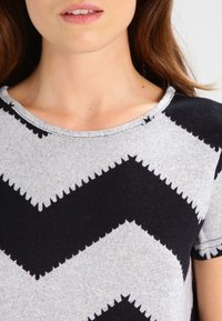 T-shirt grigio a maniche corte con motivo a zigzag nero, dotato di scollatura rotonda e texture morbida. Caratteristiche includono bordi frastagliati sui zigzag.