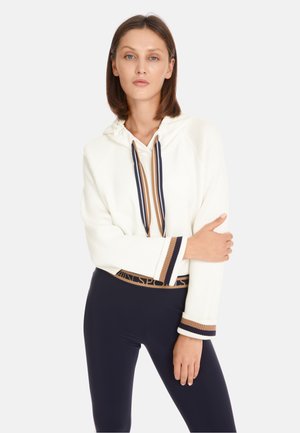 Weißer Ripp-Hoodie mit einer Kordel in Marineblau und Braun, ausgestattet mit gestreiften Bündchen und einem Bund mit Logo. Kombiniert mit Marineblauen Leggings.