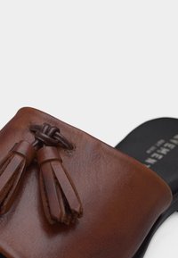 Καφέ δερμάτινο σανδάλι σλάιντ με διακοσμητικές φούντες στο πάνω μέρος και μαύρη σόλα που διακρίνεται εν μέρει στο φόντο.