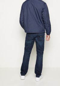 Chaqueta bomber de marina con un acabado brillante, combinada con jeans de mezclilla azul oscuro. Los jeans tienen un diseño clásico de cinco bolsillos y costuras sutiles.
