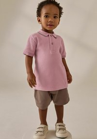 Polo shirt rosa con texture e maniche corte, abbinato a pantaloni corti marroni. Scarpe chiare con strap in Velcro. In piedi su un piedistallo circolare.