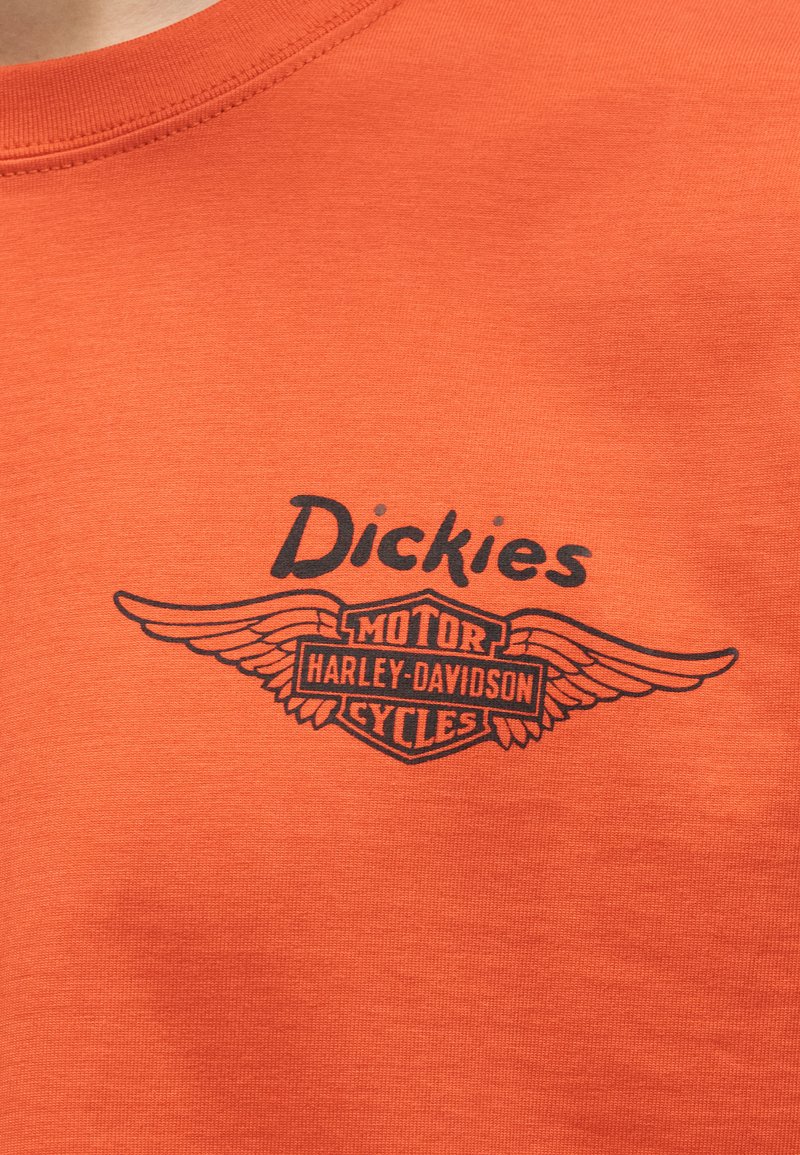 Oranges Hemd mit schwarzem "Dickies"-Text über dem Harley-Davidson Motor Cycles Flügel-Logo, das auf der Brust gedruckt ist.