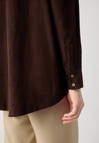 Camicia in velluto a coste marrone con vestibilità rilassata, caratterizzata da un polsino con bottoni e un orlo leggermente curvo, abbinata a pantaloni beige.