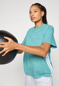 Turkos bomull t-shirt med korta ärmar, prydd med lila och vita ränder på sidorna, tillsammans med grå shorts. Håller en svart basketboll.