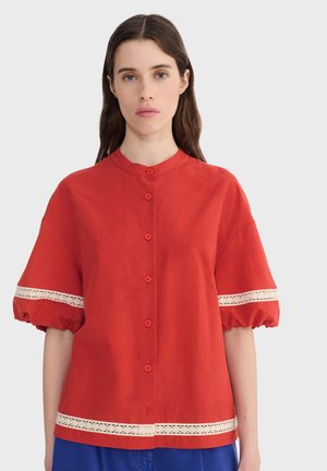 Mujer con cabello largo y castaño lleva una blusa roja abotonada con mangas cortas abullonadas y bordes de encaje blanco, combinada con pantalones azules, fondo neutral.