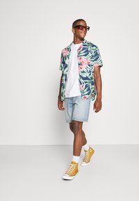 Kurzärmliges Hemd mit Blumenmuster und grünen Blättern sowie rosa Blüten, kombiniert mit Denim-Shorts und gelben High-Top-Sneakern. Klare Sonnenbrille.