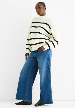 Pull en maille blanc avec des rayures noires, associé à un jean bleu à jambes larges. Des chaussures plates noires complètent la tenue.