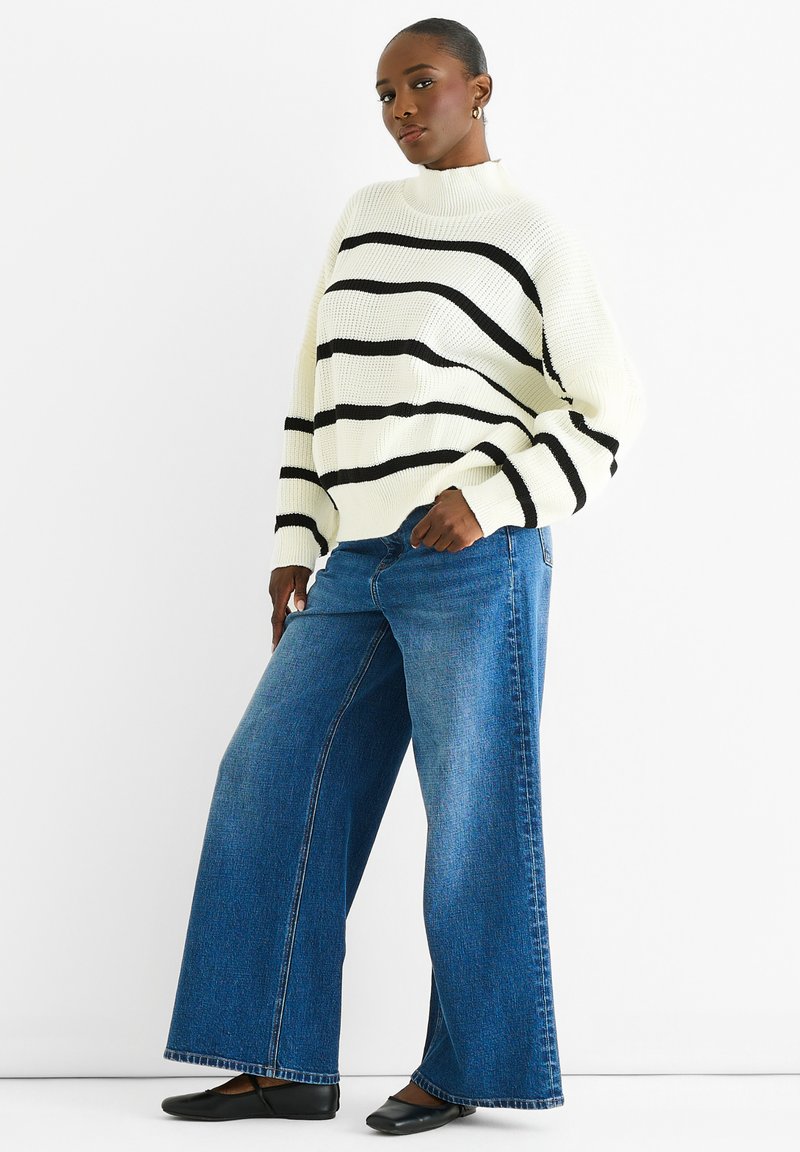 Maglione bianco lavorato a maglia con righe nere, abbinato a jeans a gamba larga blu. Scarpe basse nere completano l'outfit.